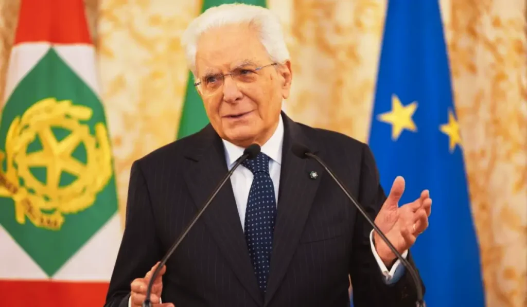 Sergio Mattarella al Quirinale durante una cerimonia istituzionale, con bandiera italiana sullo sfondo.