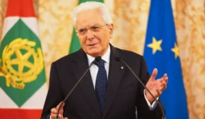 Sergio Mattarella al Quirinale durante una cerimonia istituzionale, con bandiera italiana sullo sfondo.